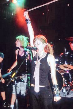 a woman in black shirt and tie standing on stage with other people behind her holding up their arms