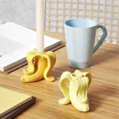 two yellow banana shaped candles next to a coffee cup on a wooden table with notebooks