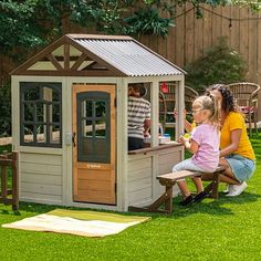 a woman and child sitting in a small wooden house on the grass near a fence