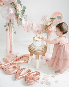 a baby girl standing in front of a cake with balloons and streamers around it