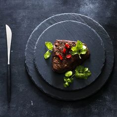 a piece of meat on a black plate next to a knife and fork with green leafy garnish