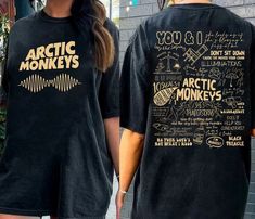 two women wearing black shirts with arctic monkeys on them and gold foil lettering that reads you & i