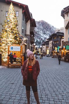 a woman standing in front of a christmas tree on a brick road next to buildings