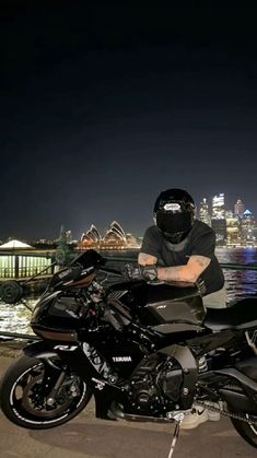 a man sitting on top of a motorcycle next to the water at night with city lights in the background