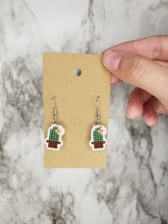 a pair of handmade earrings with green and brown beaded cactus on it, in front of a marble background