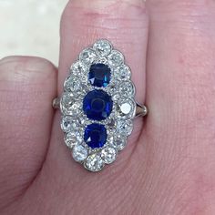 This antique ring hand-crafted in the Edwardian era features three natural sapphires in the center weighing approximately 1 carat total. A cluster of diamonds surrounds the center stone. This ring was made circa 1910 in platinum. This ring can be worn as an Engagement ring or a cocktail ring. ✦ SPECIFICATIONS: Sapphires: 1.00 Carats ✦ ENGAGEMENT RING SPECIFICATIONS: Ring Material: Platinum Stones: Blue Sapphire, Diamond ✦ WHAT COMES IN YOUR SHIPMENT: - Your Engagement Ring - Quality Ring Box - J Sapphire Diamond Rings, Dimond Ring, Antique Cushion Cut Diamond, Estate Diamond Jewelry, Edwardian Engagement Ring, Rings Antique, Edwardian Ring, Antique Diamond Rings, Sapphire And Diamond Ring