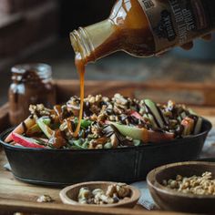 a person pouring dressing into a bowl filled with vegetables and nuts on a wooden table