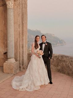 a bride and groom pose for a photo in front of an old building overlooking the ocean