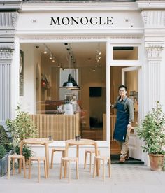 a man standing in front of a store with wooden tables and chairs around it's entrance