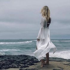 a woman standing on top of a sandy beach next to the ocean wearing a white dress