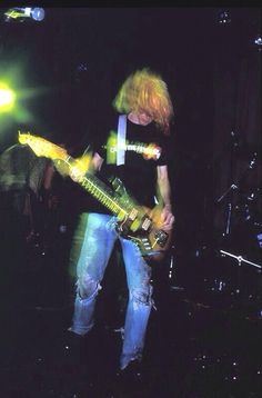 a man with long hair playing a guitar on stage in front of other people and lights