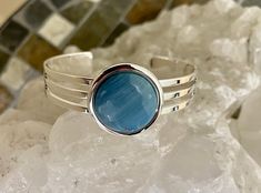 This lovely cuff bracelet is set with a spectacular Leland blue stone. The stone is 20 mm in diameter. This bracelet has a substantial silver coating that adjusts to accommodate ost wrist sizes. It does not tarnish as easily as Sterling silver does. Leland Blue is not a natural stone but rather a byproduct of smelting iron ore in the late 1800s. Blue Sterling Silver Round Cuff Bracelet, Blue Cuff Bracelet Gift, Blue Nickel-free Bangle Cuff Bracelet, Blue Round Cuff Bracelet For Formal Occasions, Adjustable Blue Gemstone Cuff Bracelet, Blue Polished Bangle Cuff Bracelet, Leland Blue Stone, Blue Cuff Bracelet, Cuff Bracelet Silver