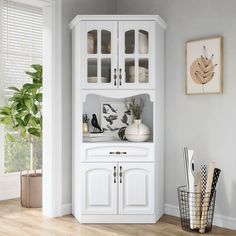 a white china cabinet with glass doors in a living room next to a potted plant