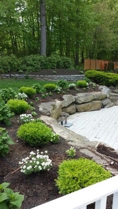 a white deck surrounded by plants and rocks in the middle of a yard with trees