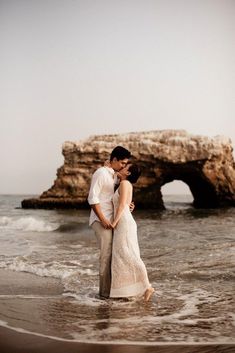 a man and woman standing in the ocean next to an old rock formation with waves crashing on them