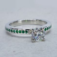 a diamond and emerald ring on a white surface with green stones in the band around it