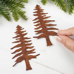 two metal christmas trees sitting on top of a white table next to evergreen branches and pine cones