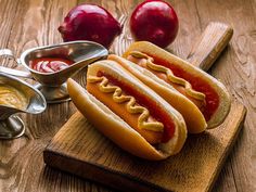 two hotdogs with ketchup and mustard on a cutting board next to apples