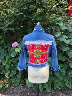 a blue jean jacket with red and green designs on it sitting on top of a white mannequin