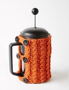 an orange knitted coffee pot holder with black handles and buttons on the top, sitting on a white surface