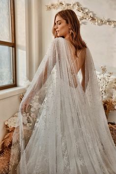 a woman in a white wedding dress with a long veil on her head is standing next to a window