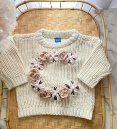 a white sweater with pink flowers on it sitting in a wicker basket next to a flower arrangement