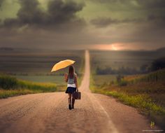 a woman walking down a road holding an umbrella over her head while the sun is setting