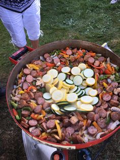 a big pot filled with lots of food on top of a grass covered field next to a person