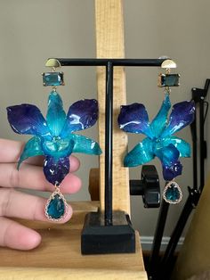 These stunning earrings feature real blue orchids delicately preserved in resin, creating a unique and eye-catching piece of jewelry. The orchids are complemented by gold-plated half moon studs and elegant blue glass charms, adding a touch of sophistication to the design. **Details - Real blue and purple orchids preserved in resin - Gold-plated half moon studs - Blue glass charms - Drop length: 3-4 inches Each pair of earrings is handmade with care, making them a perfect gift for nature lovers or anyone who appreciates beautifully crafted jewelry. Add a pop of color and elegance to your outfit with these exquisite orchid earrings! The colors and shapes of each orchid might be slightly different as they are made of real orchid flowers.This is a precious item that deserves your utmost attent Blue And Purple Orchids, Orchid Earrings, Orchid Flowers, Blue Orchids, Moon Studs, Purple Orchids, Glass Charms, Jewelry Lookbook, Stunning Earrings