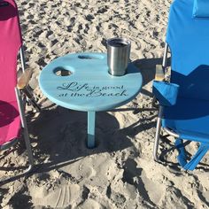 two beach chairs and a table with a cup on it sitting in the sand at the beach