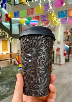 a hand holding up a black cup in front of a colorful wall with lights hanging from it