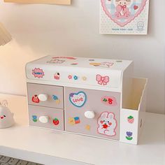 a white desk topped with a toy chest next to a lamp and pictures on the wall