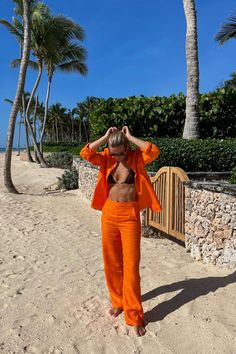 a woman in an orange outfit standing on the beach with palm trees and blue sky behind her