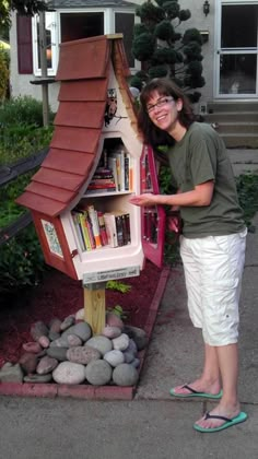 a woman standing next to a doll house made out of bookshelves and rocks