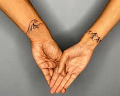 two hands holding each other with tattoos on their wrists and fingers, both showing the same direction