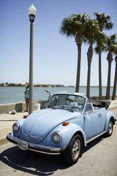 an old blue car parked on the side of the road near some palm trees and water