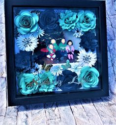 a shadow box with blue paper flowers and two dolls in the center, sitting on a wooden surface