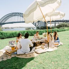 a group of people sitting around a picnic table with an umbrella over it and the sydney harbour bridge in the background