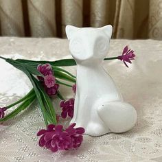 a white cat figurine sitting next to purple flowers