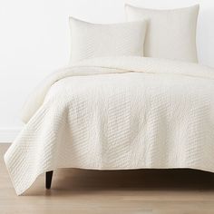 a white bedspread with two pillows on top of it and a wooden floor