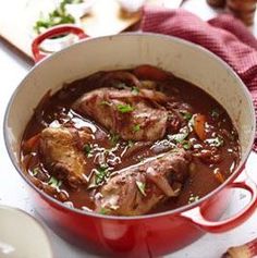 a red pot filled with meat and vegetables on top of a white counter next to bread