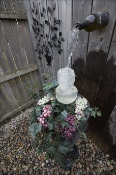 a water fountain in the middle of a garden filled with flowers and greenery next to a wooden fence