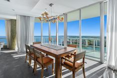 a dining room table with chairs and a chandelier in front of large windows overlooking the ocean