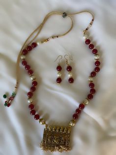 The Nashili necklace set is made with maroon and and pearl beads, with a plaque pendant depicting two handmaids fanning Ganesha in traditional Indian metalwork. The subtle details of the metalwork elevate the piece, from the borders around the plaque to the setting of the pearl beads to the structure of the necklace itself. The adjustable backing allows for some adjustments to the length of the necklace, although it is meant to hang long past the chest. Traditional Festive Jewelry Set With Pearl Pendant, Festive Traditional Jewelry Set With Pearl Pendant, Traditional Pearl Pendant Jewelry Set For Celebration, Red Pearl Necklace Temple Jewelry For Festivals, Red Pearl Temple Jewelry Necklace For Festivals, Red Temple Jewelry Pearl Necklace For Festivals, Red Pearl Necklace For Festive Gift, Red Pearl Necklace Gift For Festive Occasions, Festive Red Pearl Necklace As Gift