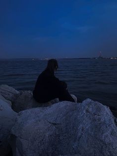 a person sitting on top of a rock next to the ocean at night with a full moon in the sky