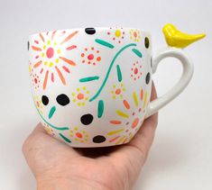 a hand holding a painted coffee cup with a yellow bird on the handle and sprinkles