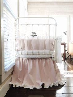 a white crib with pink ruffled bedding in a room next to a window