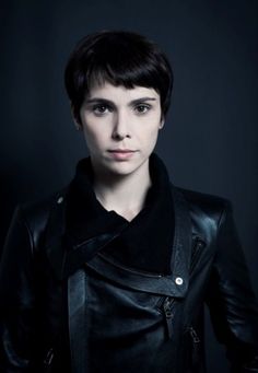 a woman with short hair wearing a black leather jacket and looking at the camera while standing in front of a dark background