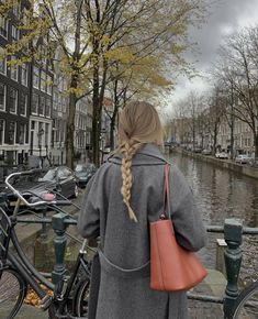 a woman standing next to a bike on the side of a river with buildings in the background