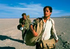 two men are walking in the desert carrying their belongings and one is carrying a backpack